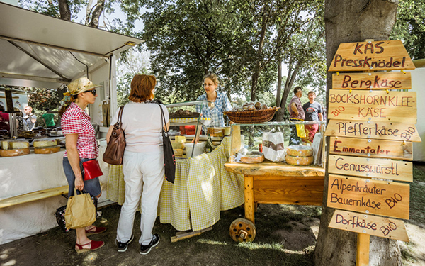3 Personen vor einem Marktstand beim Genussfestival im Stadtpark.. Verkauft werden verschiedene Käse-Produkte.