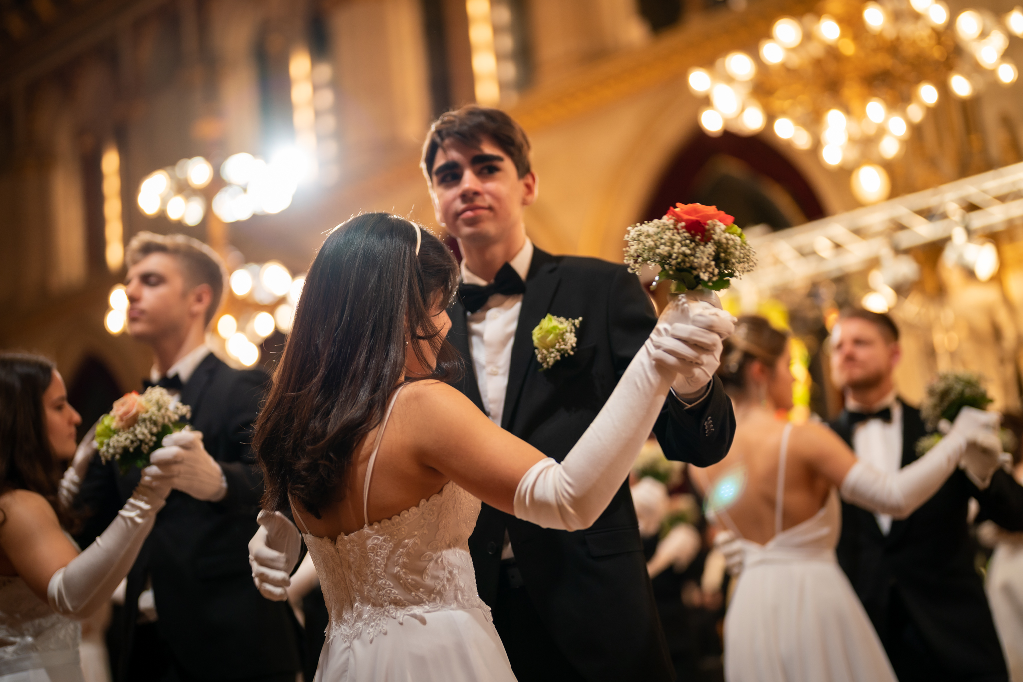 Walzer tanzende Paare mit einem kleinen Blumenstrauß in der Hand. Die Frauen tragen weiße, lange Kleider mit Handschuhen, die Männer Anzüge.