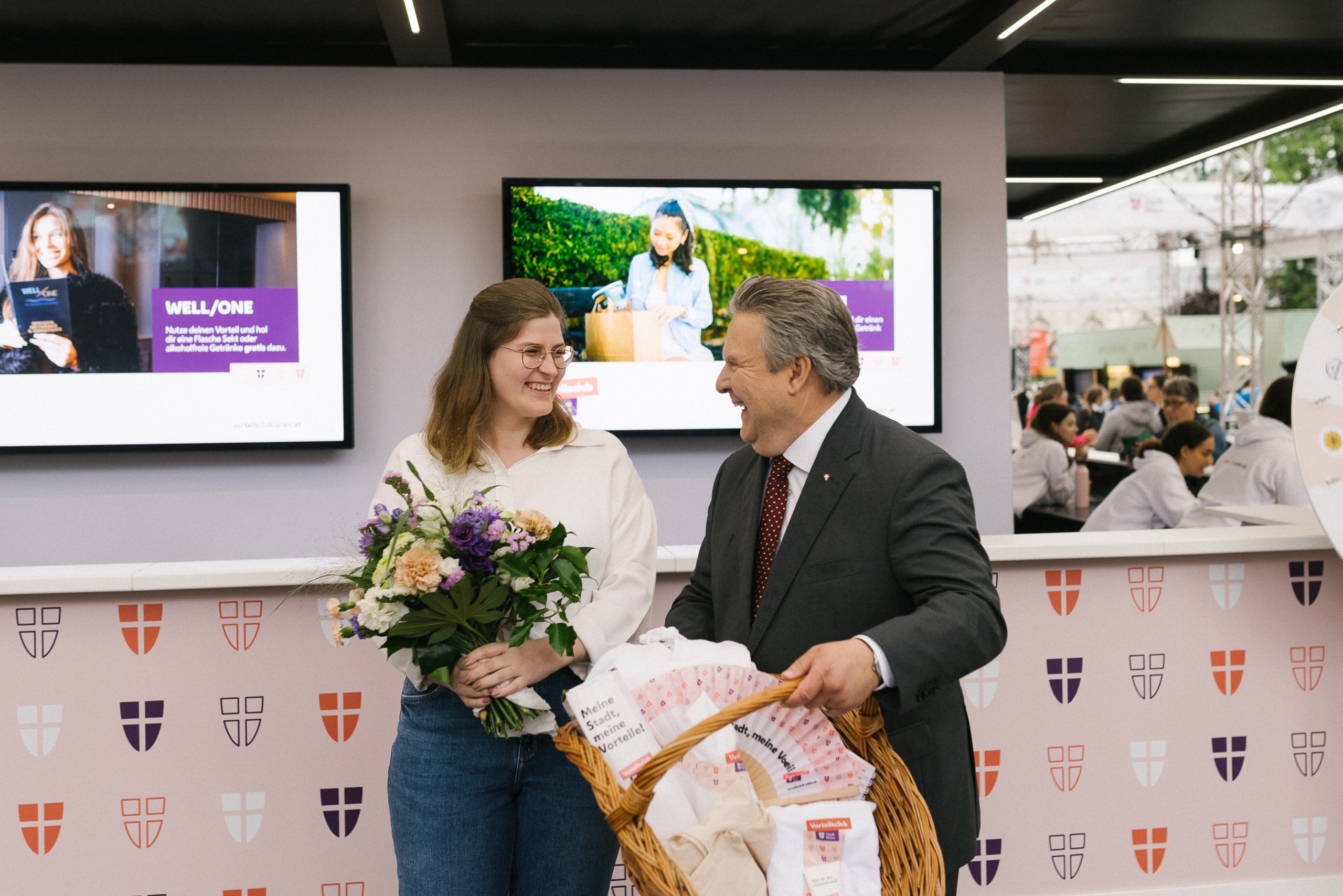Eine Frau und Bürgermeister Michael Ludwig mit einem Blumenstrauß und Geschenkkorb vor einem Vorteilsclub Stand.