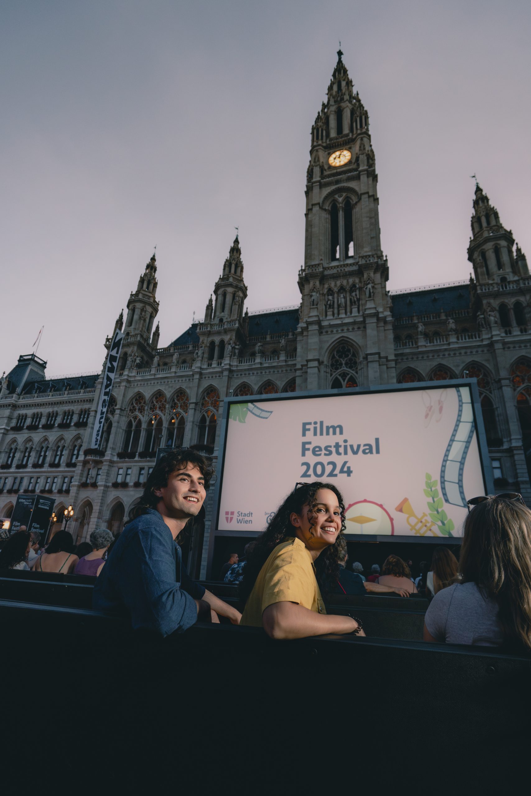 Zwei Personen sitzen auf den Bänken vor der Leinwand beim Film Festival und drehen sich über ihre linke Schulter nach hinten. Die Frau trägt ein gelbes Oberteil und der Mann ein blaues. Auf der Leinwand steht auf einem hellrosa Hintergrund "Film Festival 2024".