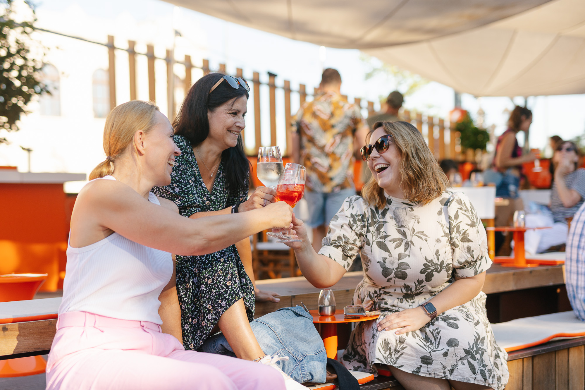 3 Frauen sitzen auf einer Lounge und stoßen mit ihren orangen Getränken an und lächeln sich dabei an.
