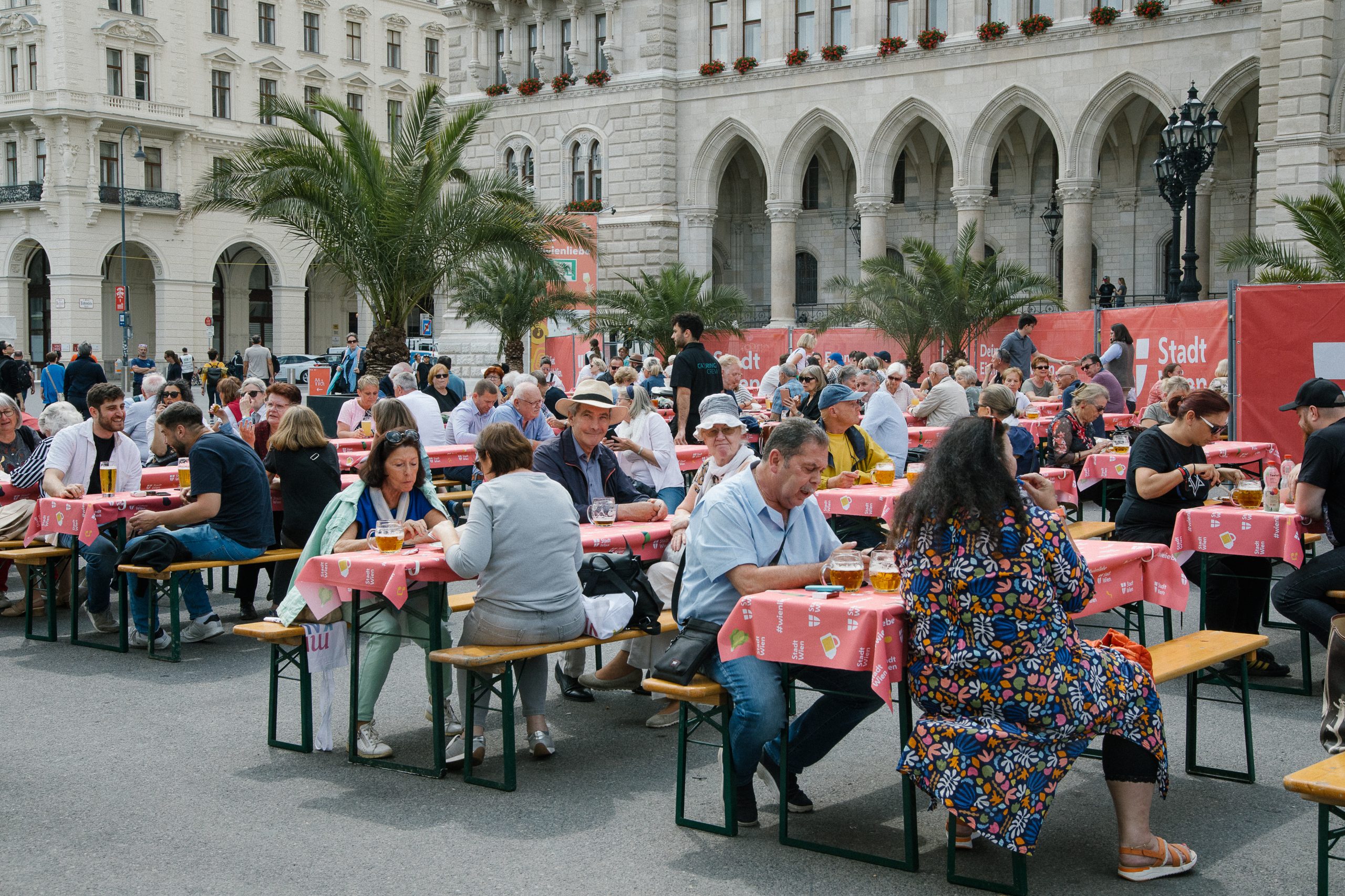 © stadt wien marketing / Studio Wey Personen beim Wienliebe Festival auf Bierzeltgarnituren beim Essen und Trinken. Im Hintergrund ist ein Teil des Rathaus zu sehen.