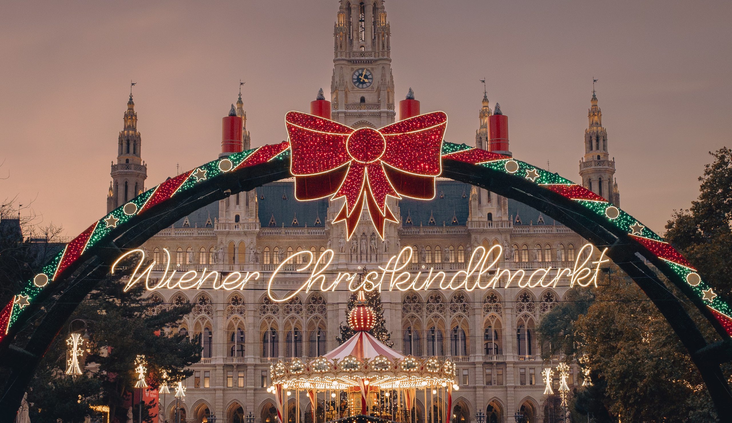 Frontale Sicht auf das Rathaus und einem großen Eingangsbogen mit dem Schriftzug "Wiener Christkindlmarkt". Zu sehen ist noch ein beleuchtetes Karussell.