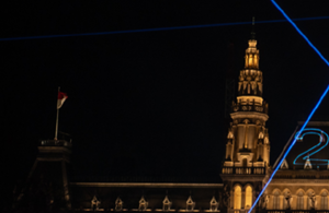 Teil eines aufgesplitteten Fotos des Rathaus. Auf diesem ist die linke Seite des Dachs mit einem kleinen Turm und dem Dach bei Nacht zusehen. Das Rathaus ist erleuchtet und es sind Laser zu sehen. Die Zahl 2 ist mit einem Laser auf das Rathaus projiziert.