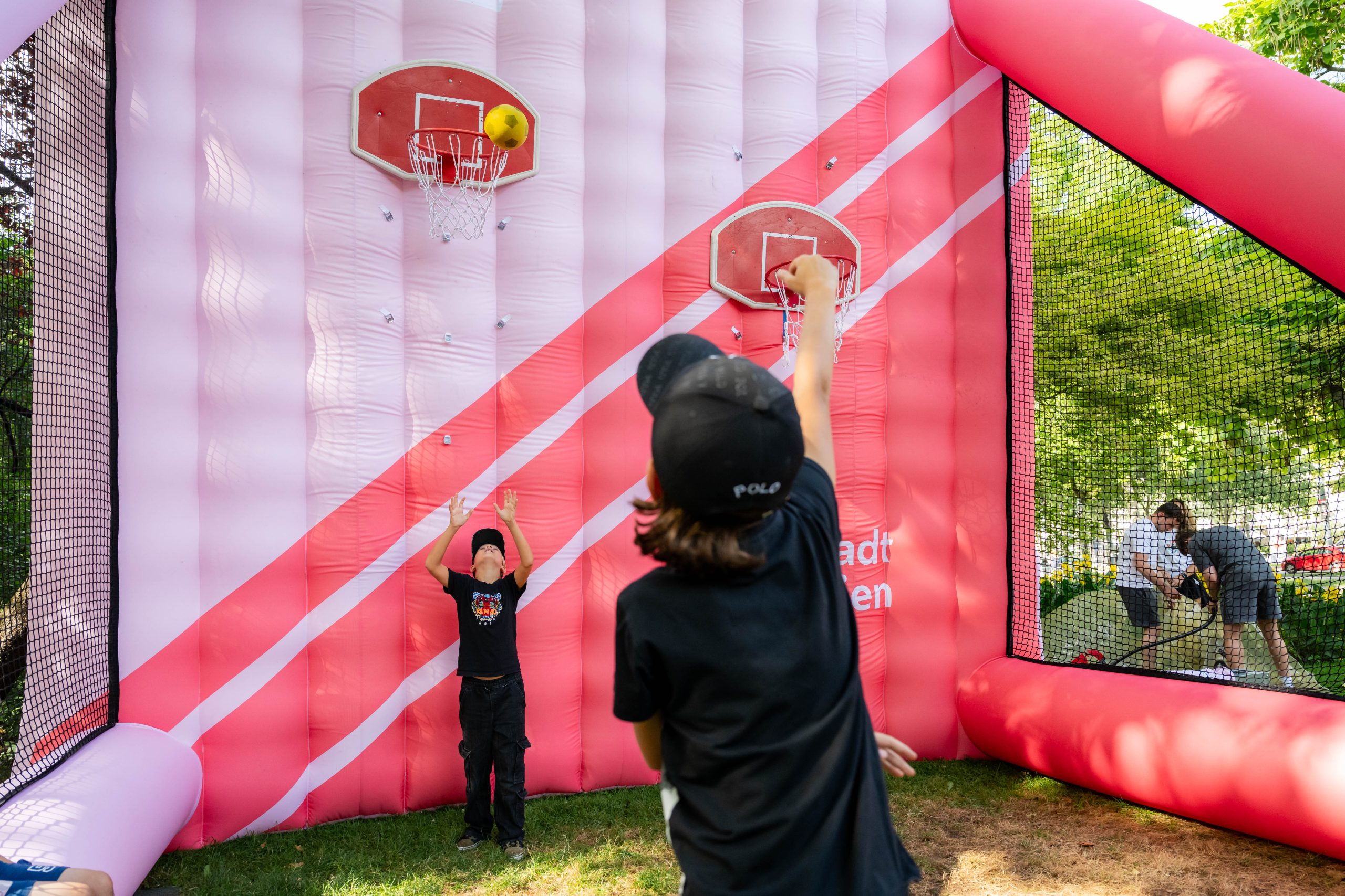 Zwei Buben in schwarzer Kleidung stehen vor einer aufblasbaren Wand, an der ein Basketballkorb befestigt ist. Ein Junge wirft einen Basketball, der andere steht mit ausgestreckten Armen unter dem Korb.