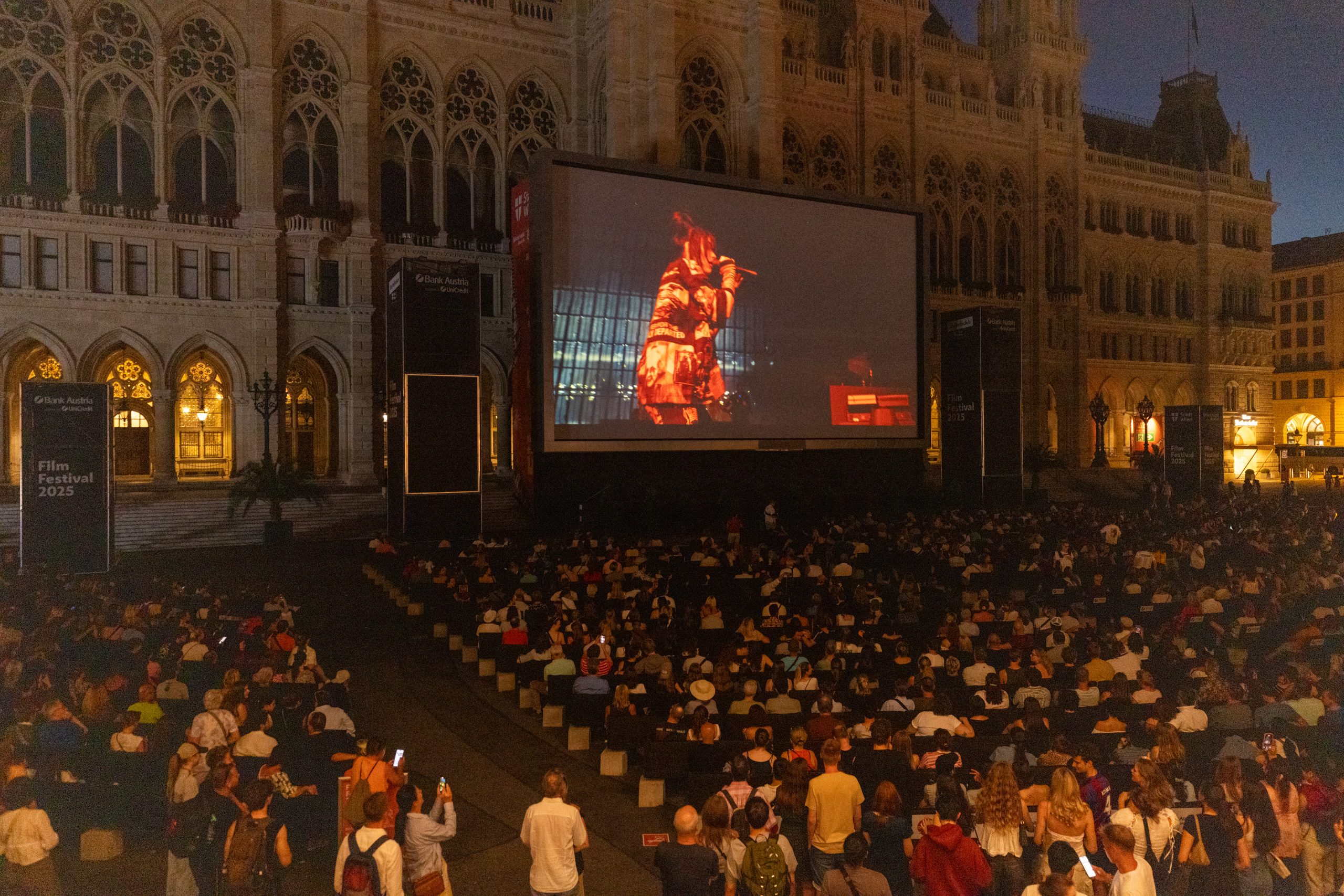 © stadt wien marketing / Georg Krewenka Blick auf die große Leinwand beim Filmfestival. Es ist ein Teil des Rathaus zu sehen. Vor der Leinwand sitzt eine große Menschenmenge. Gezeigt wird ein Konzert von Billie Eilish.