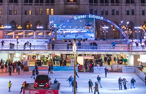 Teil eines aufgesplitteten Fotos des Rathaus. Auf diesem ist der Rathausplatz mit einer großen Eisfläche und Eisläufer*innen zu sehen. In der Mitte der Fläche steht ein rotes Werbeauto auf einem Podest.