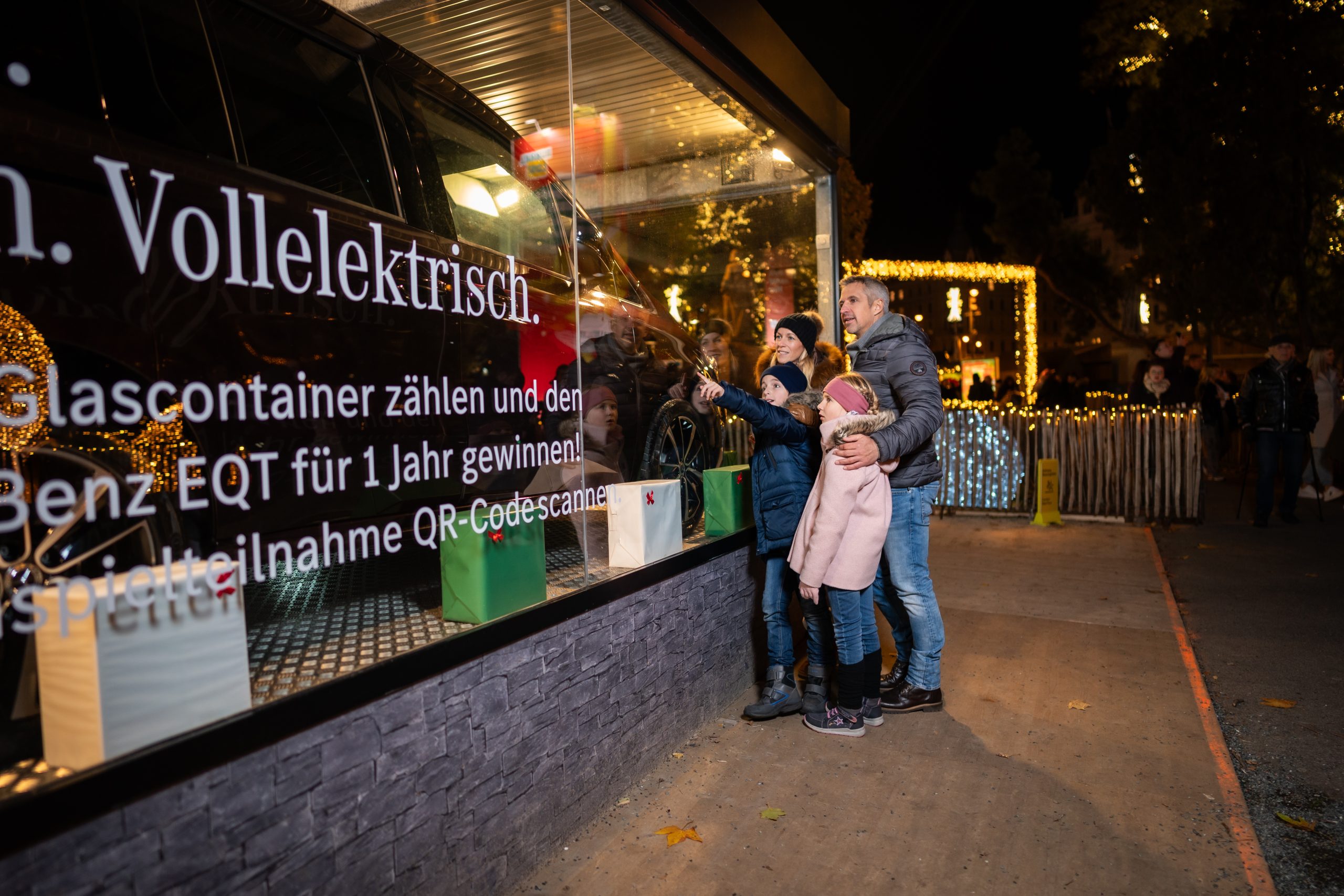 © stadt wien marketing / David Bohmann Ein Mercedes Bus in einem Glascontainer. Vor dem Bus im Container stehen kleine Weihnachtsgeschenke. Eine vierköpfige Familie steht vor dem Container und schaut das Auto an.