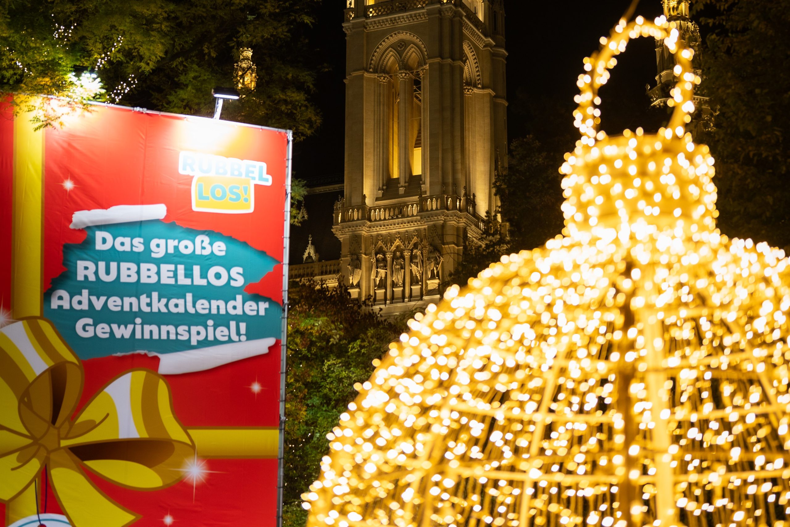 © stadt wien marketing / David Bohmann Rechts im Bild ist ein überdimensional großer Rubbellos Kalender der Österreichischen Lotterien. In der Mitte im Hintergrund das erleuchtete Rathaus und links eine große, leuchtende Weihnachtskugel.