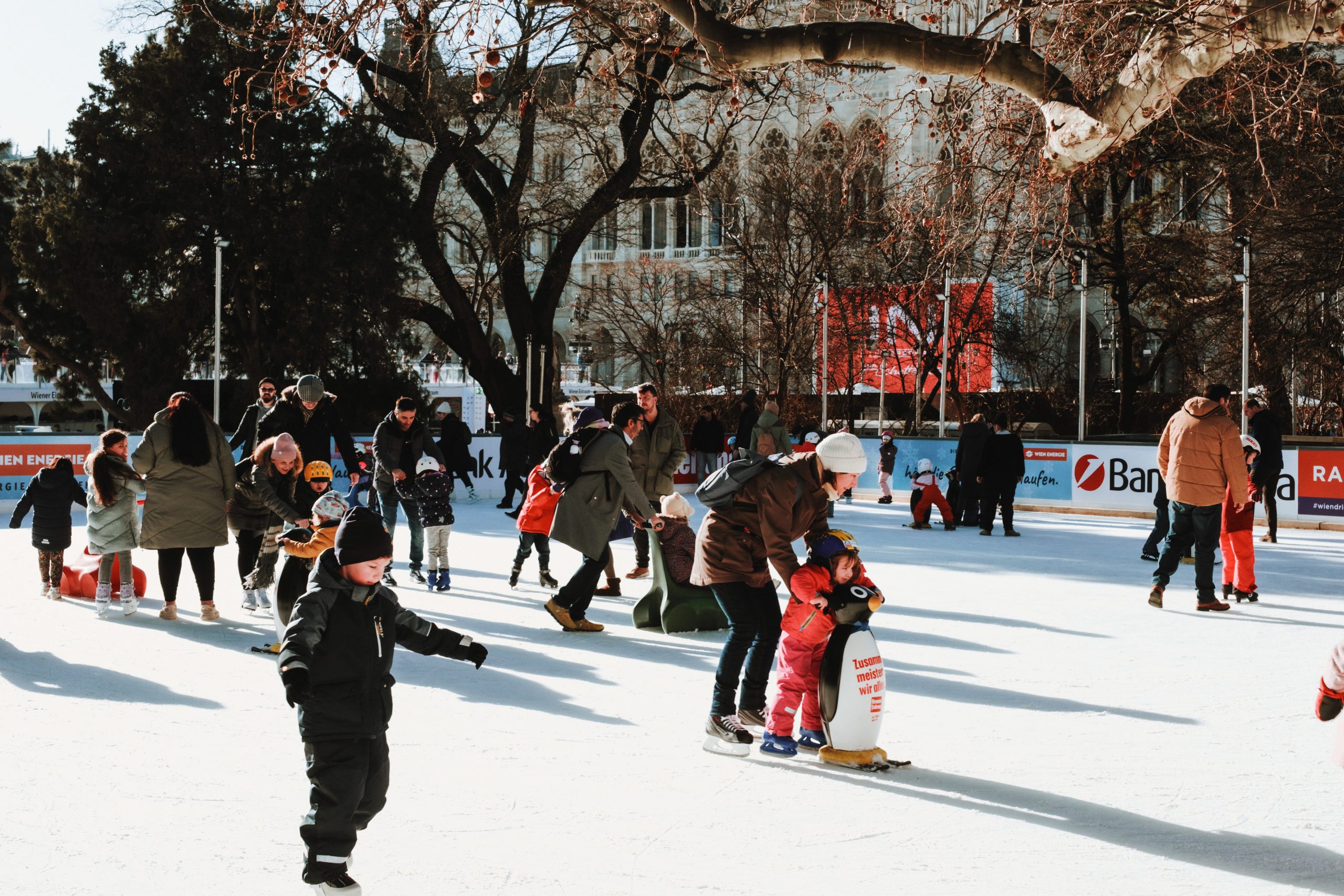 Auf dem Foto ist eine der Eisflächen vom Wiener Eistraum bei strahlend blauen Himmel gezeigt. Viele Menschen, vor allem Kinder, sind zusehen. Im Hintergrund befindet sich das Rathaus.
