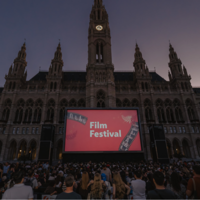 Design ohne Titel bendaufnahme des Wiener Rathauses während des Film Festival. Vor dem beleuchteten Rathaus steht eine große rote Leinwand mit der Aufschrift „Film Festival“. Davor sitzen zahlreiche Menschen auf den Zuschauerplätzen und genießen die Atmosphäre am Abend.