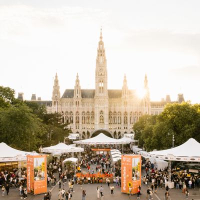 Design ohne Titel Das Wienliebe Festival vor dem Wiener Rathausplatz ist zu sehen. Menschen gehen über den Platz. Ein Skyliner, verschiedene Stände und Aufsteller zum Wienliebe Festival befinden sich auf dem Gelände.