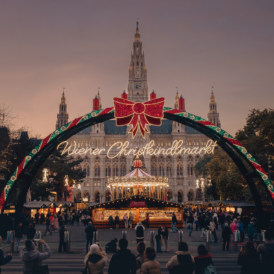 Wiener Christkindlmarkt Das Wiener Rathaus im Hintergrund des Christkindlmarkts am Rathausplatz. Vor dem Rathaus befinden sich festlich dekorierte Stände, ein Karussell und zahlreiche Menschen, die den Weihnachtsmarkt besuchen.